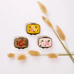 Three small embroidered patchwork cases with a cream-colored fabric exterior and colorful floral and berry embroidery, arranged on a white background with dried grass stems.