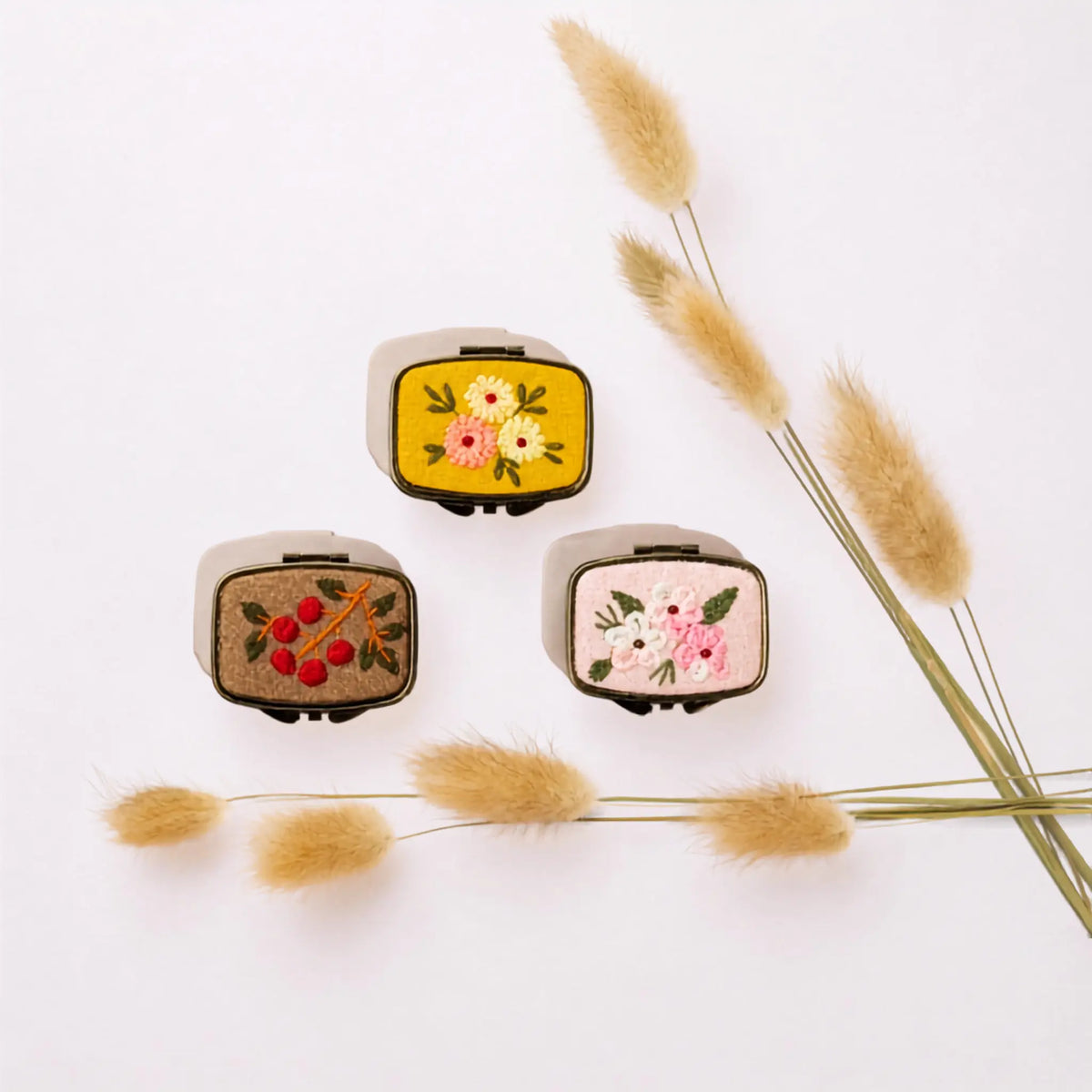 Three small embroidered patchwork cases with a cream-colored fabric exterior and colorful floral and berry embroidery, arranged on a white background with dried grass stems.
