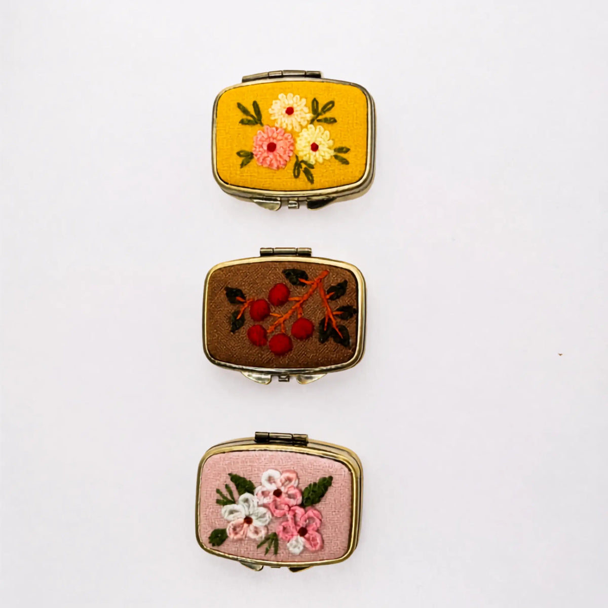 Three vintage-style rectangular compact mirrors with rounded corners, each featuring different embroidered floral designs on colored fabric covers, arranged vertically on a plain white background.