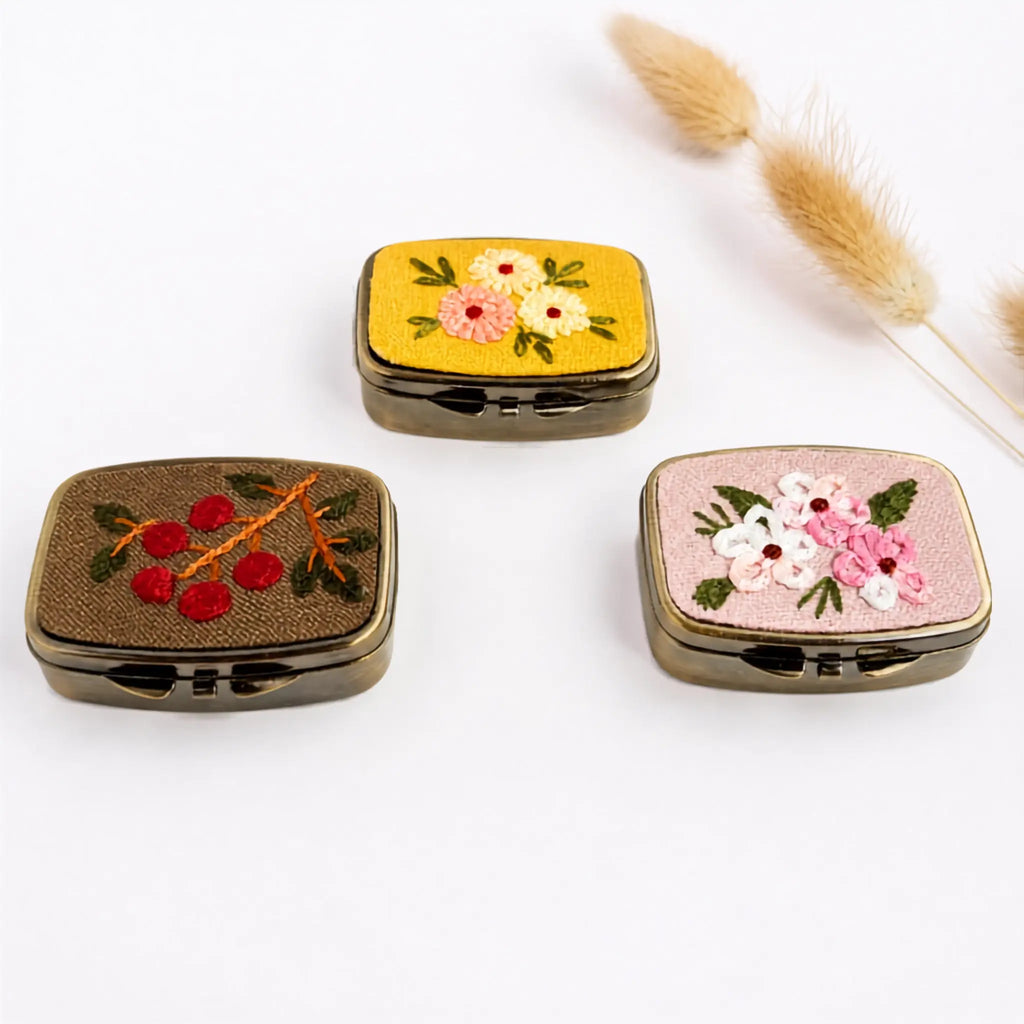 Three small embroidered metal compact cases arranged on a white background, with dried bunny tail grass in the upper right. The cases have dark bronze-toned edges and lids featuring floral and berry embroidery: one yellow with pink and white flowers, one brown with red berries, and one pink with white and pink flowers.