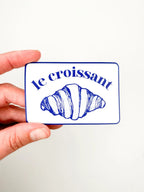 Card with a croissant illustration and 'le croissant' text held by a hand against a white background