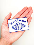 Hand holding a card with 'le croissant' and a croissant illustration on a white background