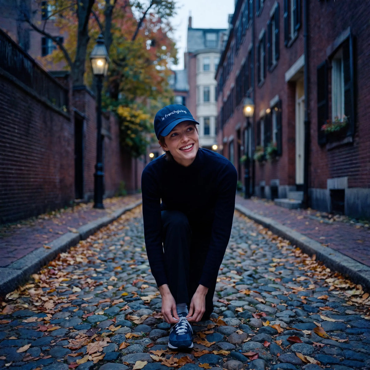 A woman crouches on a cobblestone street lined with brick buildings and fallen leaves, tying the laces of her running shoe. She wears a dark navy long-sleeve top, dark pants, a navy cap with white text, and a blue-and-white running shoe.
