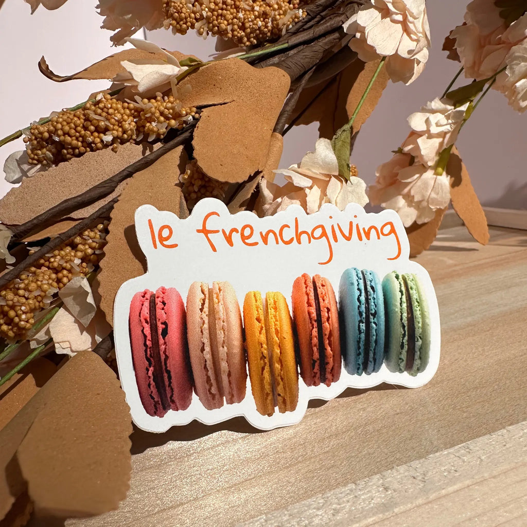 A die-cut sticker featuring the phrase 'le frenchiving' in orange script above six colorful macarons, displayed on a wooden surface with dried flowers in the background.