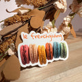 A die-cut sticker featuring the phrase 'le frenchiving' in orange script above six colorful macarons, displayed on a wooden surface with dried flowers in the background.
