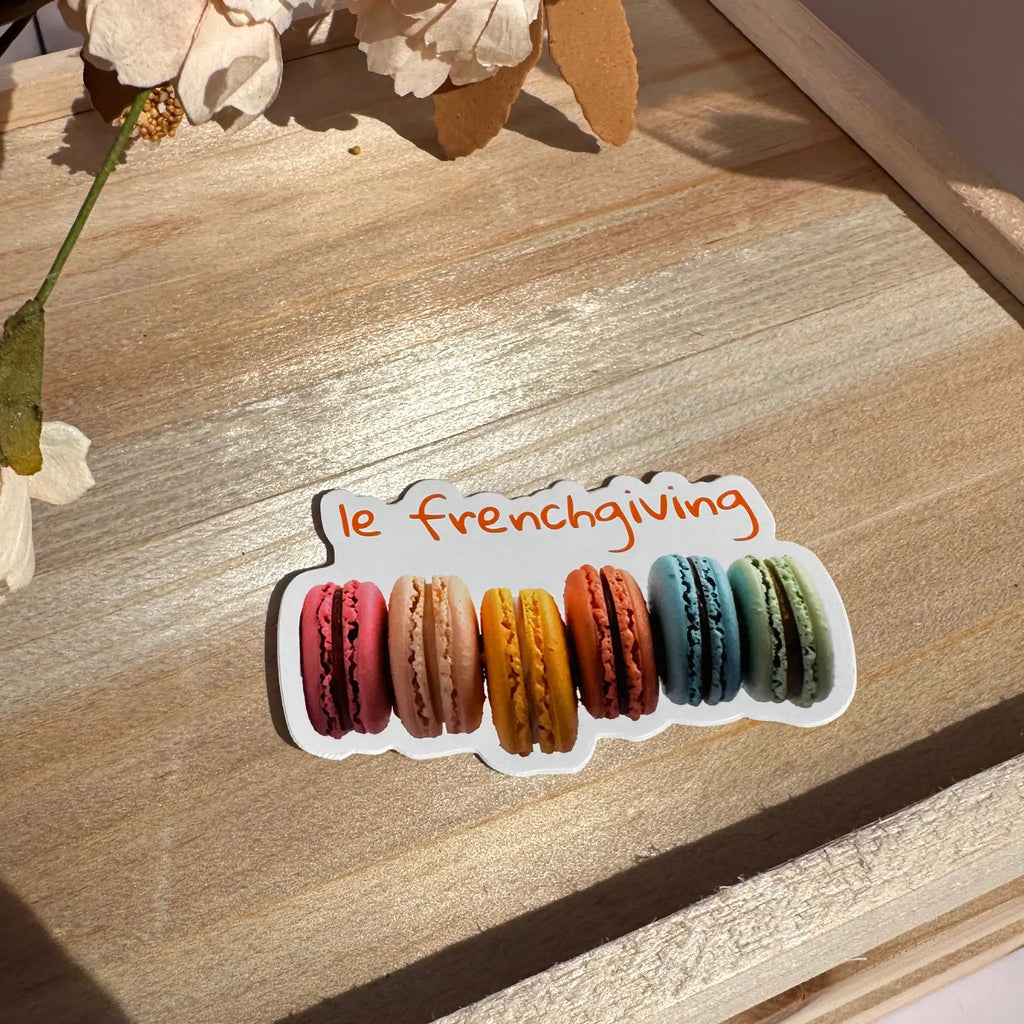 A die-cut sticker featuring six colorful French macarons with the phrase 'le frenchiving' in orange script above them, placed on a wooden surface with dried flowers in the background.