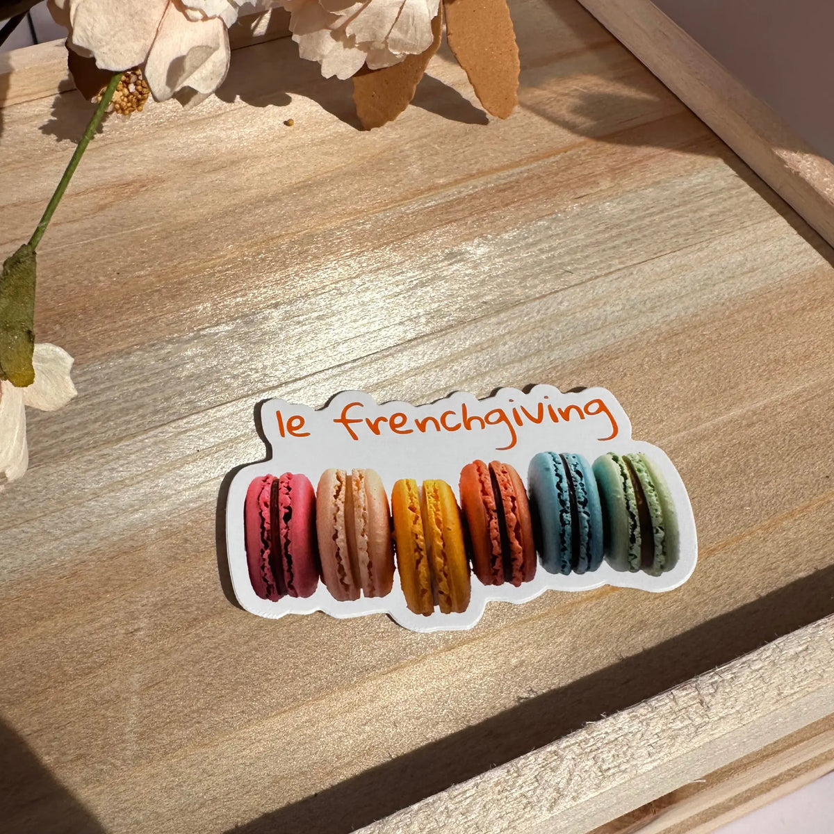 A die-cut sticker featuring six colorful French macarons with the phrase 'le frenchiving' in orange script above them, placed on a wooden surface with dried flowers in the background.