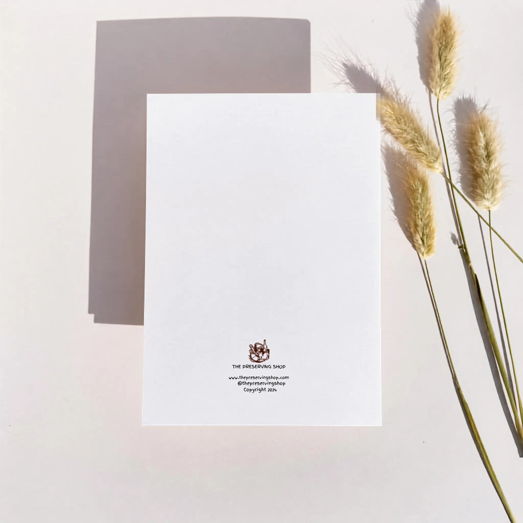 A white rectangular card or paper sheet with a small printed logo and text near the bottom center, photographed on a light neutral background with dried grass stems along the right side and a soft shadow behind the card.