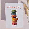 A white greeting card with the phrase 'le frenchiving' in orange script above a vertical stack of colorful macarons, styled on a light pink background with dried grass stems.