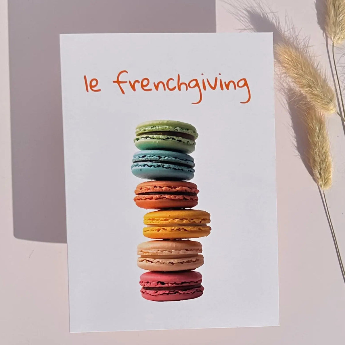 A white greeting card with the phrase 'le frenchiving' in orange script above a vertical stack of colorful macarons, styled on a light pink background with dried grass stems.
