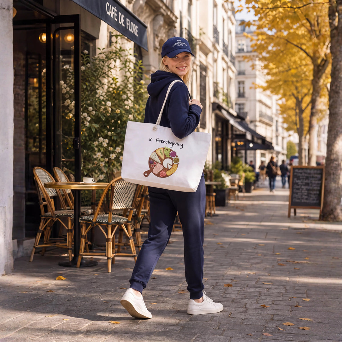 Woman in navy sweat suit walking down the street carrying a white tote bag with a charcuterie board on it with white text "le frenchgiving"