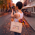 A person standing on an autumn street takes a photo while wearing a white tank top and carrying a beige canvas tote bag with the text 'le friendsgiving' and small food illustrations. The bag also contains flowers and a loaf of bread.
