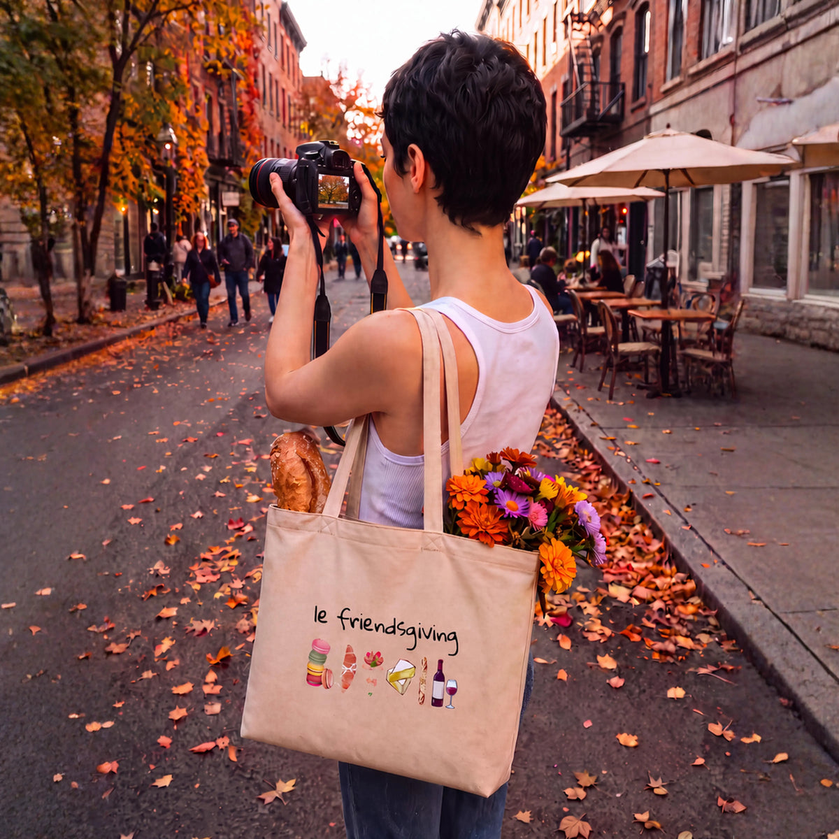 A person standing on an autumn street takes a photo while wearing a white tank top and carrying a beige canvas tote bag with the text 'le friendsgiving' and small food illustrations. The bag also contains flowers and a loaf of bread.