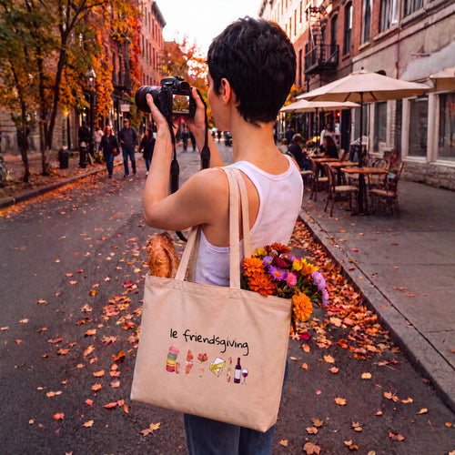 A person standing on an autumn street takes a photo while wearing a white tank top and carrying a beige canvas tote bag with the text 'le friendsgiving' and small food illustrations. The bag also contains flowers and a loaf of bread.