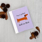 A lavender greeting card with a white envelope, featuring an orange dachshund illustration and the text 'long on gratitude short on stress', styled on a light gray textured surface with dried brown leaves and seed pods.