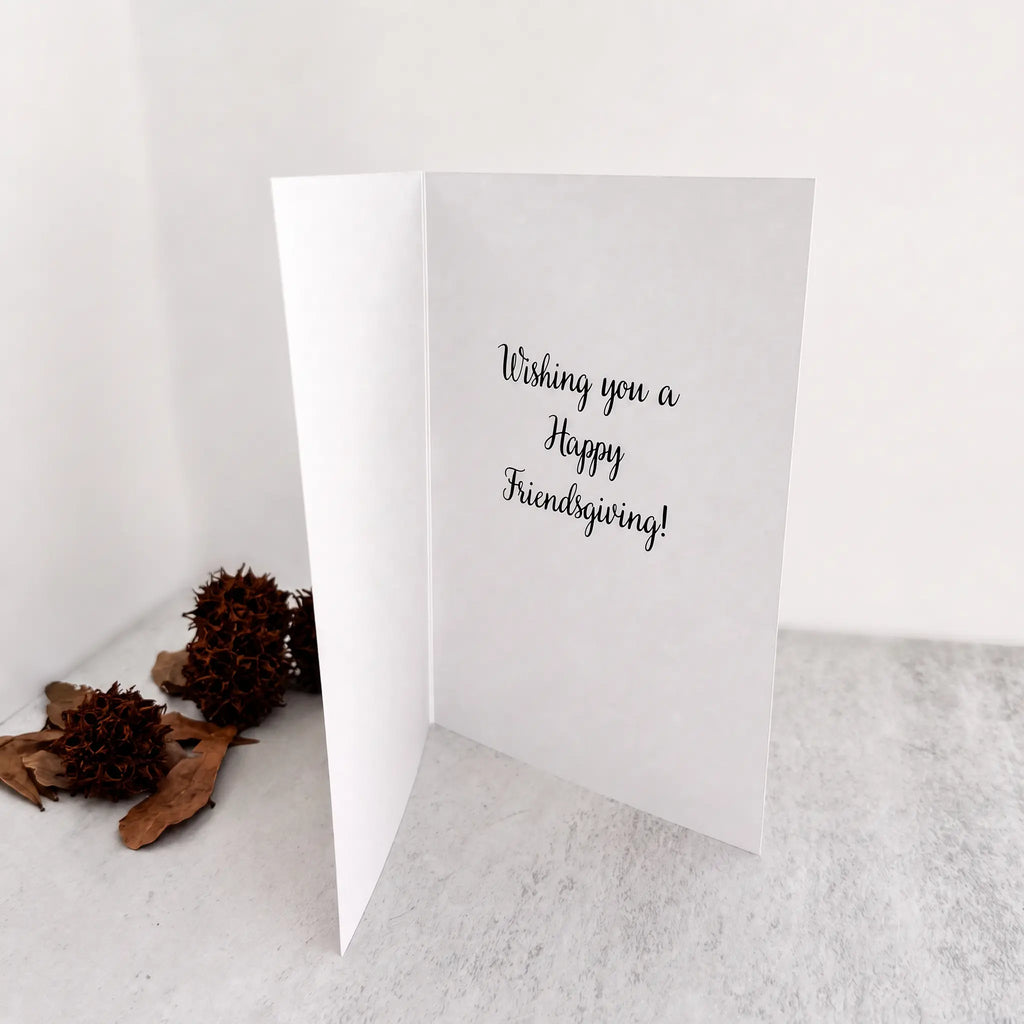 A white folded greeting card standing open on a light surface, with the handwritten-style text 'Wishing you a Happy Friendsgiving!' printed on the inside. Two brown dried flower buds and leaves sit to the left against a plain white background.