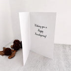A white folded greeting card standing open on a light surface, with the handwritten-style text 'Wishing you a Happy Friendsgiving!' printed on the inside. Two brown dried flower buds and leaves sit to the left against a plain white background.
