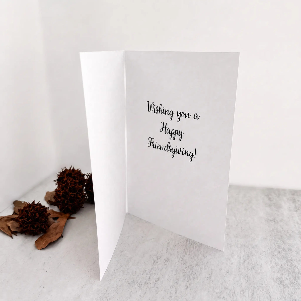 A white folded greeting card standing open on a light surface, with the handwritten-style text 'Wishing you a Happy Friendsgiving!' printed on the inside. Two brown dried flower buds and leaves sit to the left against a plain white background.