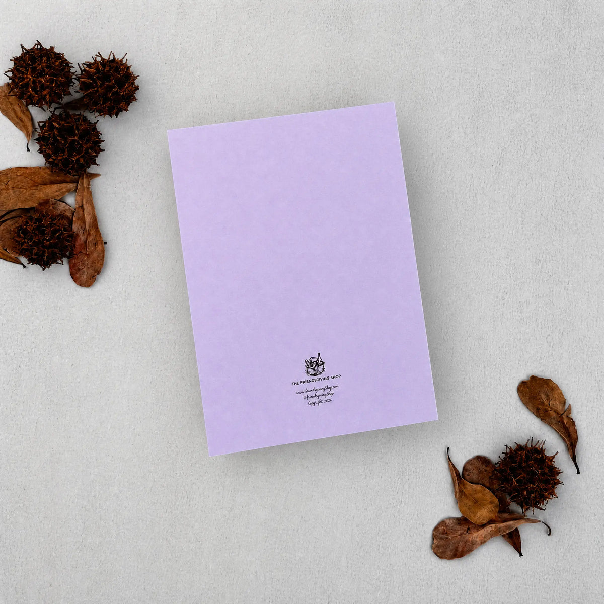A lavender purple letterhead card with a small black crest and text at the bottom, styled flat-lay on a textured light gray background with dried brown leaves and seed pods.
