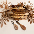 A wooden leaf-shaped serving board or tray displayed on a light wood crate, surrounded by dried floral arrangements, with two engraved wooden serving utensils in front on a white background.