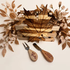 A wooden leaf-shaped serving board or tray displayed on a light wood crate, surrounded by dried floral arrangements, with two engraved wooden serving utensils in front on a white background.