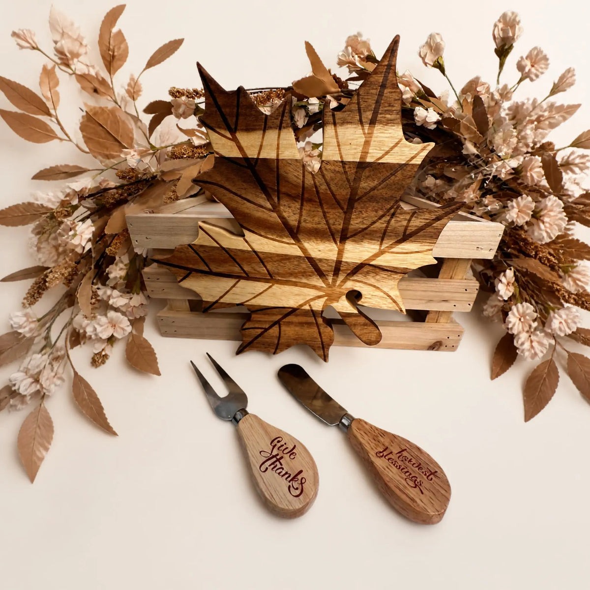 A wooden leaf-shaped serving board or tray displayed on a light wood crate, surrounded by dried floral arrangements, with two engraved wooden serving utensils in front on a white background.