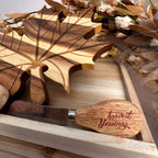 A wooden-handled cheese knife with the words 'harvest blessings' engraved on the handle, resting on a wooden tray alongside matching engraved wooden leaf-shaped pieces and dried floral decor.