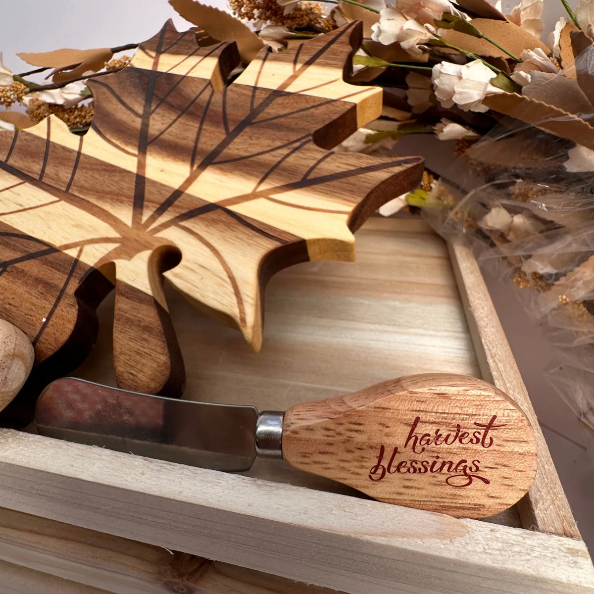 A wooden-handled cheese knife with the words 'harvest blessings' engraved on the handle, resting on a wooden tray alongside matching engraved wooden leaf-shaped pieces and dried floral decor.
