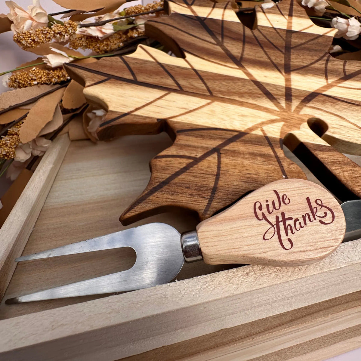 A wooden serving utensil set in a light wood gift box, featuring a leaf-shaped wooden board engraved with leaf veins and a matching wooden-handled serving fork engraved with 'Give Thanks' on the handle.