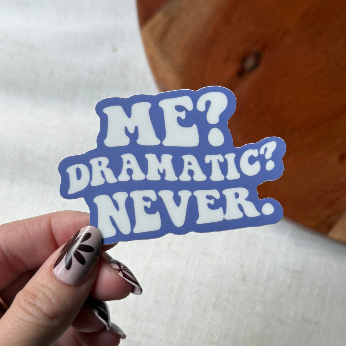 Hand holding a blue and white sticker with 'ME? DRAMATIC? NEVER.' text on a wooden surface.