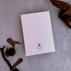 A white rectangular card or letterhead with a small black crest and text at the bottom, photographed from above on a light gray textured surface with dried leaves and driftwood props.