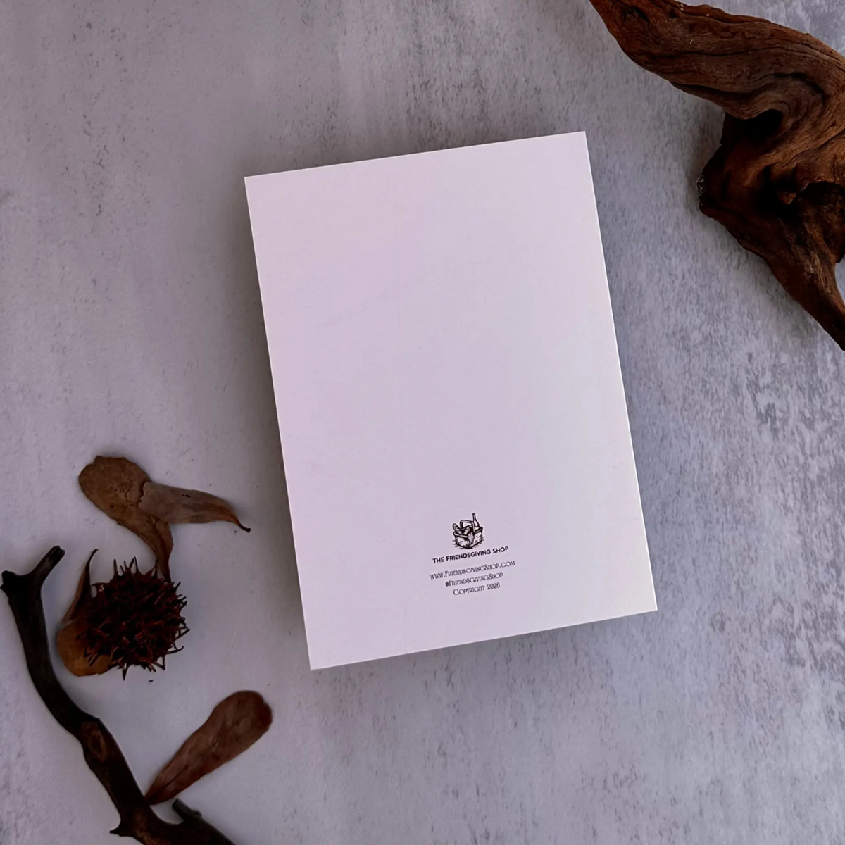 A white rectangular card or letterhead with a small black crest and text at the bottom, photographed from above on a light gray textured surface with dried leaves and driftwood props.