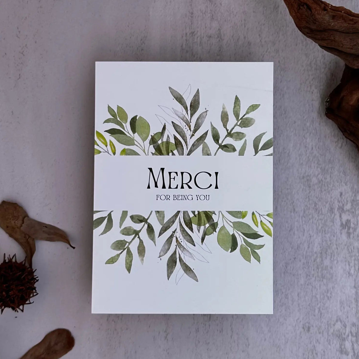 A white rectangular card or box with green leaf illustrations and the text 'MERCI FOR BEING YOU' in the center, arranged on a light gray textured surface with dried botanical props and driftwood.