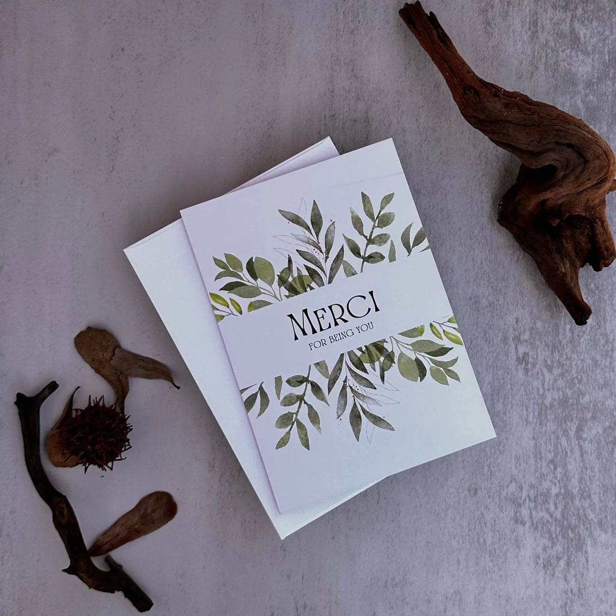 A white wedding thank-you card with green leaf illustrations and the text 'MERCI FOR BEING YOU' stacked on top of a white envelope, styled on a textured gray surface with driftwood props.