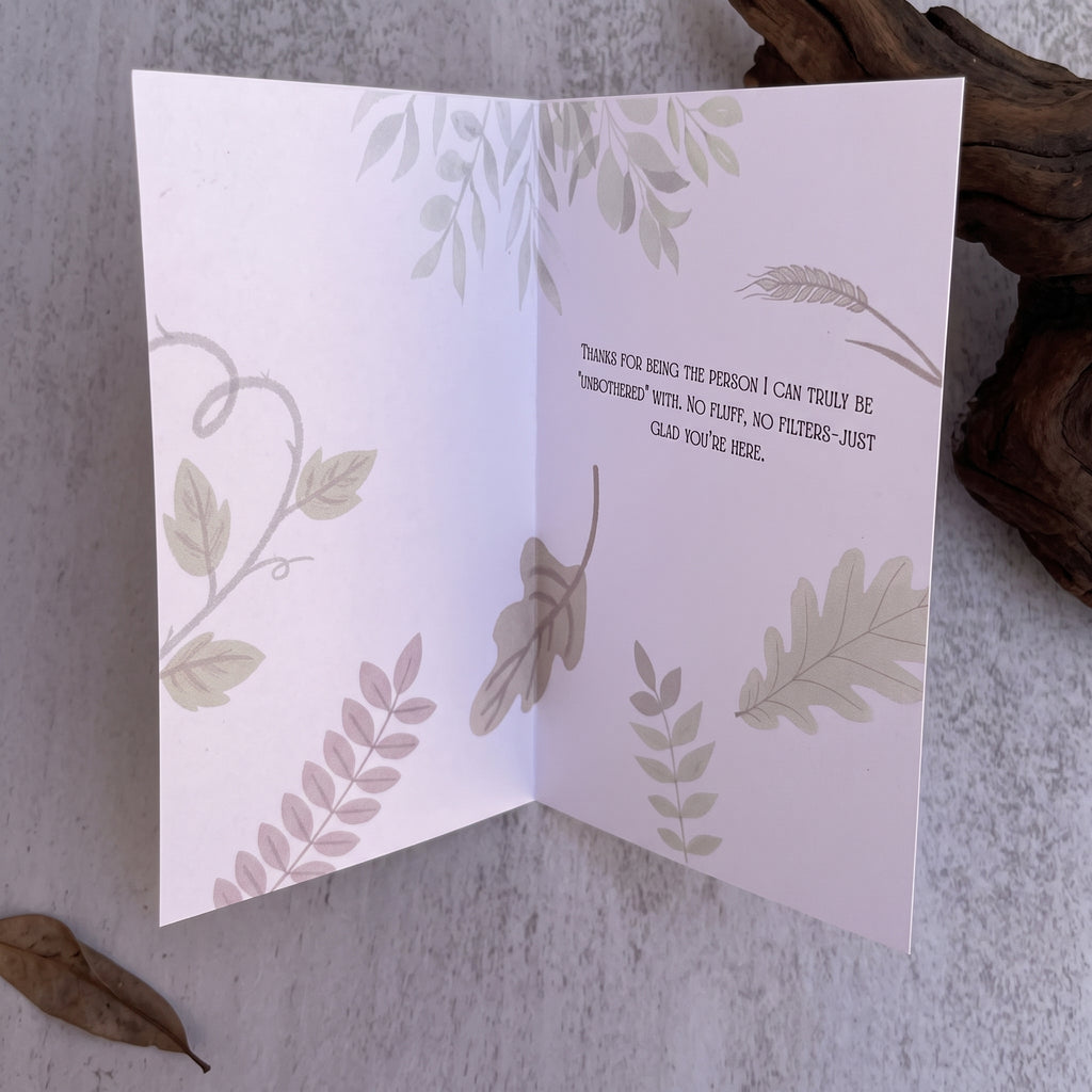 A folded greeting card displayed open on a textured light gray surface, with botanical illustrations in muted sage green, blush pink, and taupe, plus a printed message on the inside. Decorative dried leaves and driftwood are placed around it.