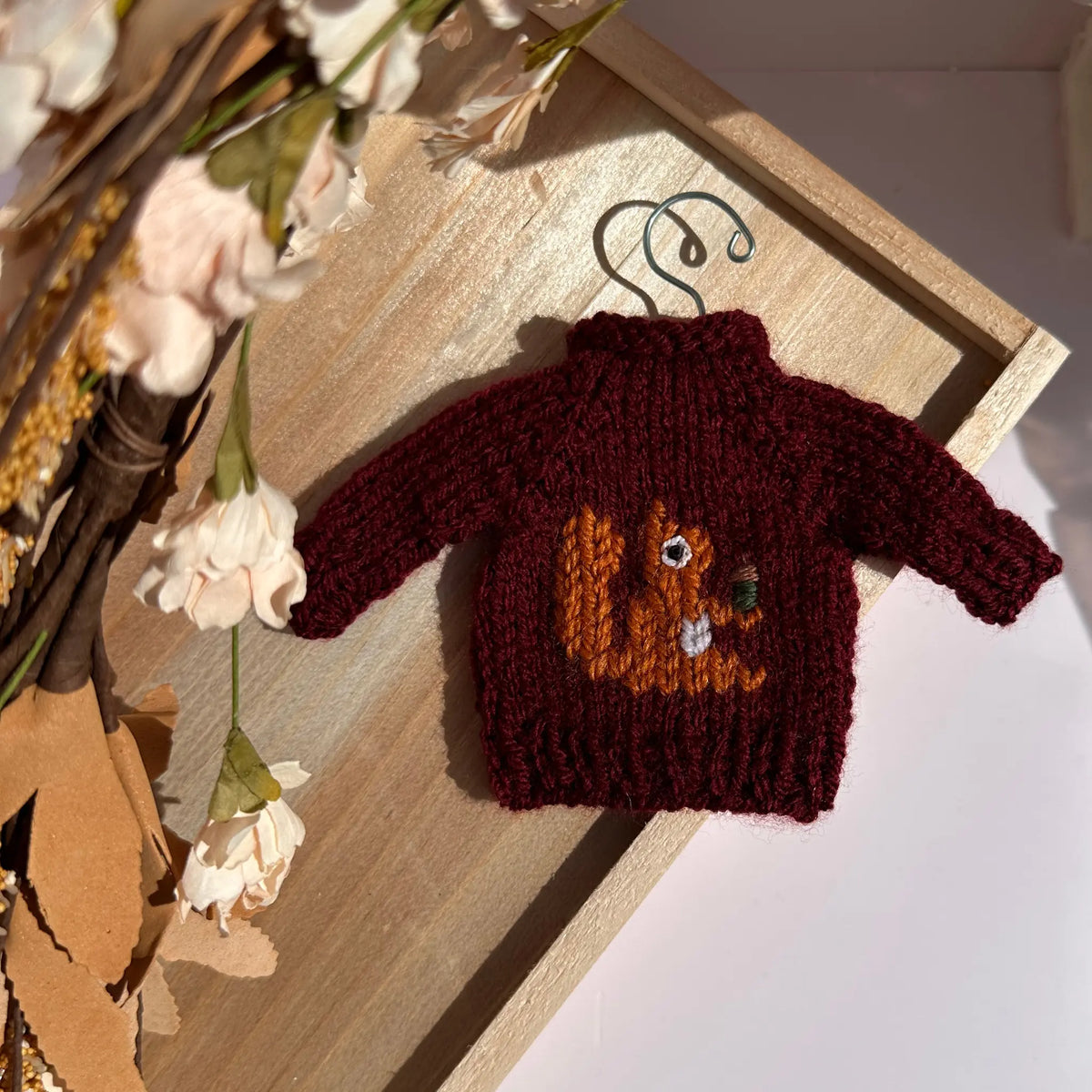 A miniature burgundy knit sweater with a pumpkin face design, displayed on a wooden tray with dried flowers.