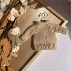 A small beige knitted sweater displayed on a metal hanger, resting on a wooden tray with dried flowers and leaves around it.