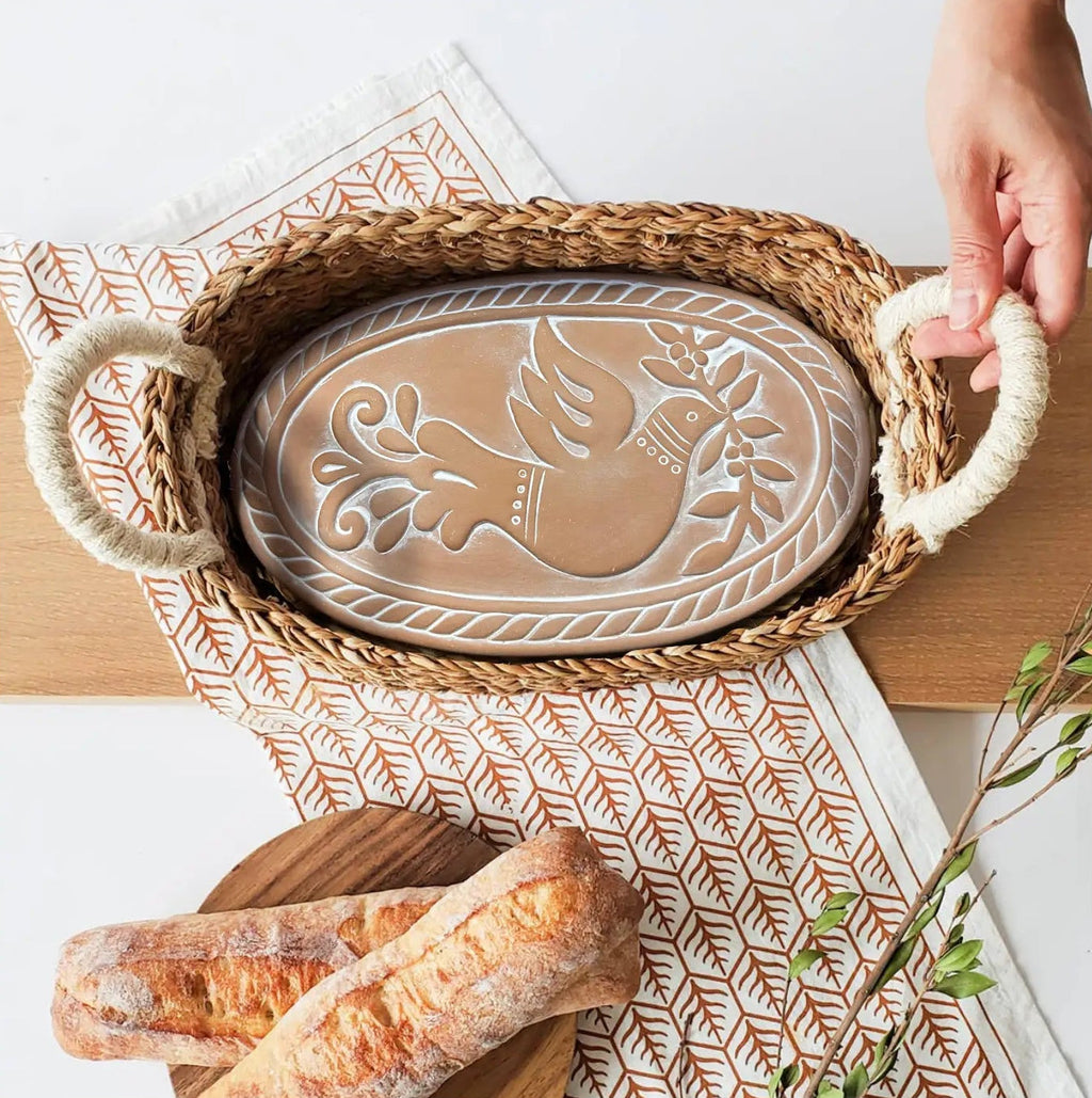 An oval woven basket with rope handles holds a carved ceramic plaque featuring a bird design. The scene is styled on a light wood surface with a patterned runner, bread on a wooden board, and a hand holding one of the basket handles.