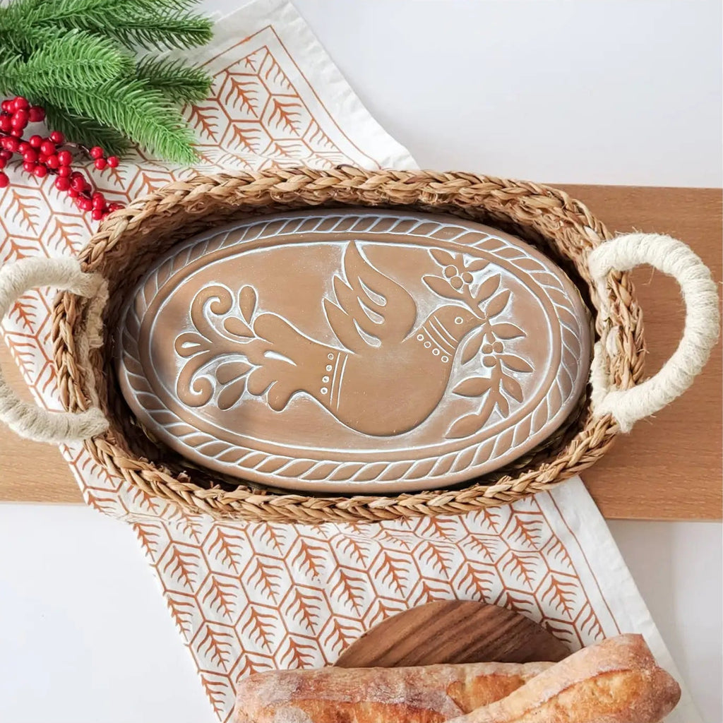 An oval woven tray with rope handles and a raised rooster motif in the center, styled on a patterned runner with holiday greenery and red berries.