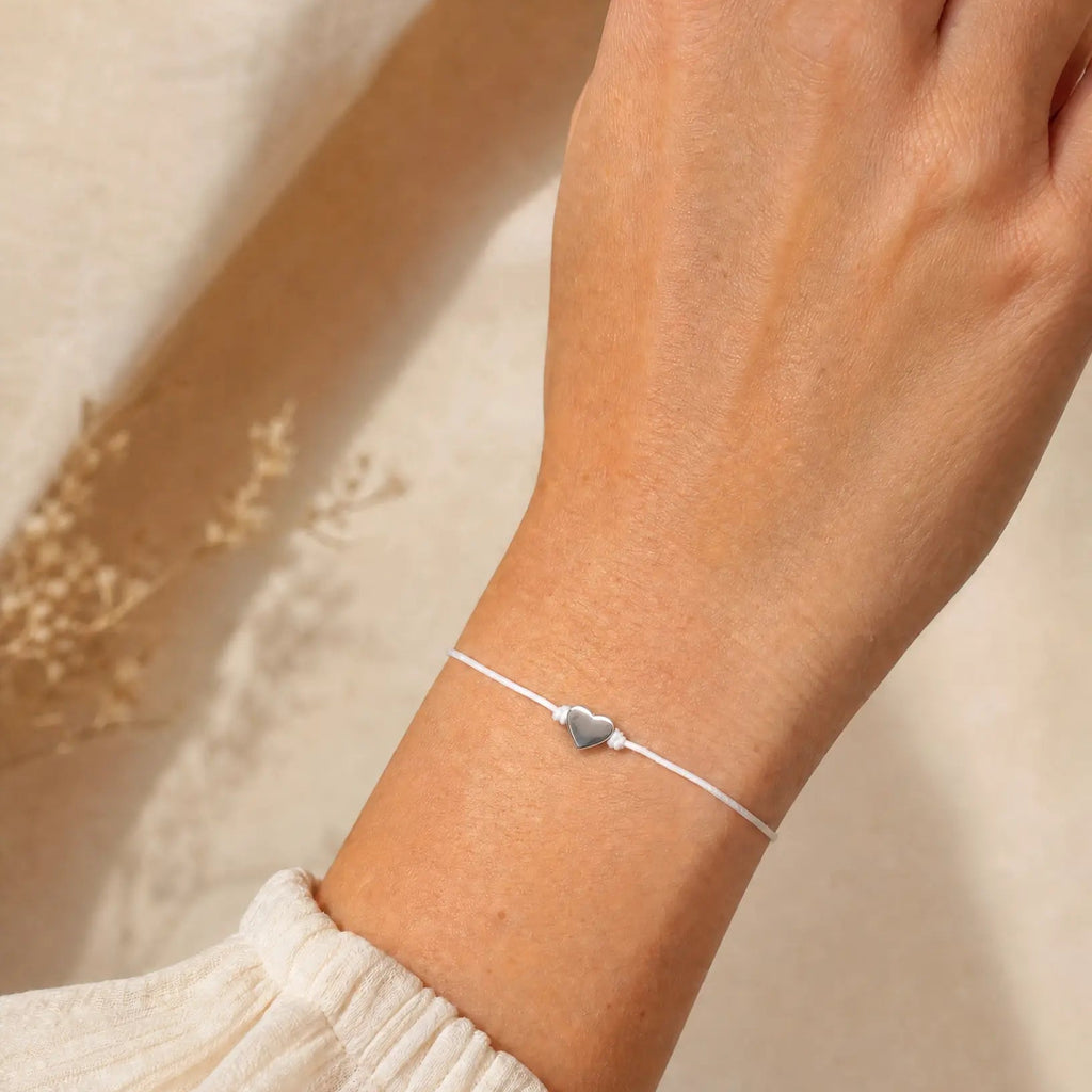 A delicate white cord bracelet with a small silver heart charm, worn on a person's wrist against a warm beige background.