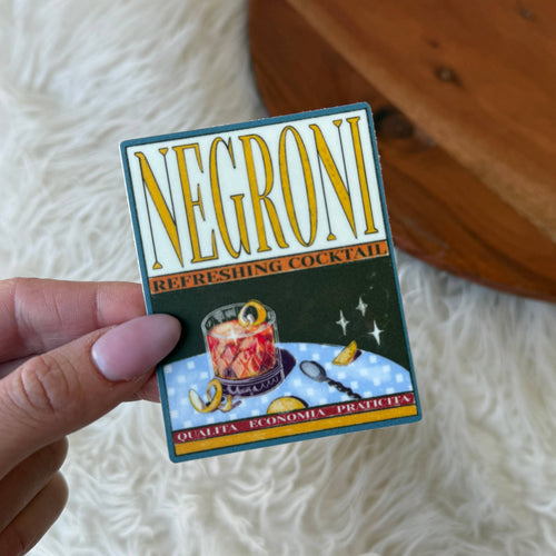 A hand with pink manicured nails holds a square vintage-style cocktail card featuring the brand name 'NEGRONI' in large yellow letters. The card has an illustrated cocktail glass, lemon slices, and decorative text, photographed against a white faux fur surface with a wooden surface in the background.