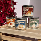 Four metal scented candles in cylindrical cans are displayed on a light wood shelf. The top can is labeled 'baked apple pie' and the three cans in front are labeled 'pumpkin chai', 'shortbread cookie', and 'matcha latte'.