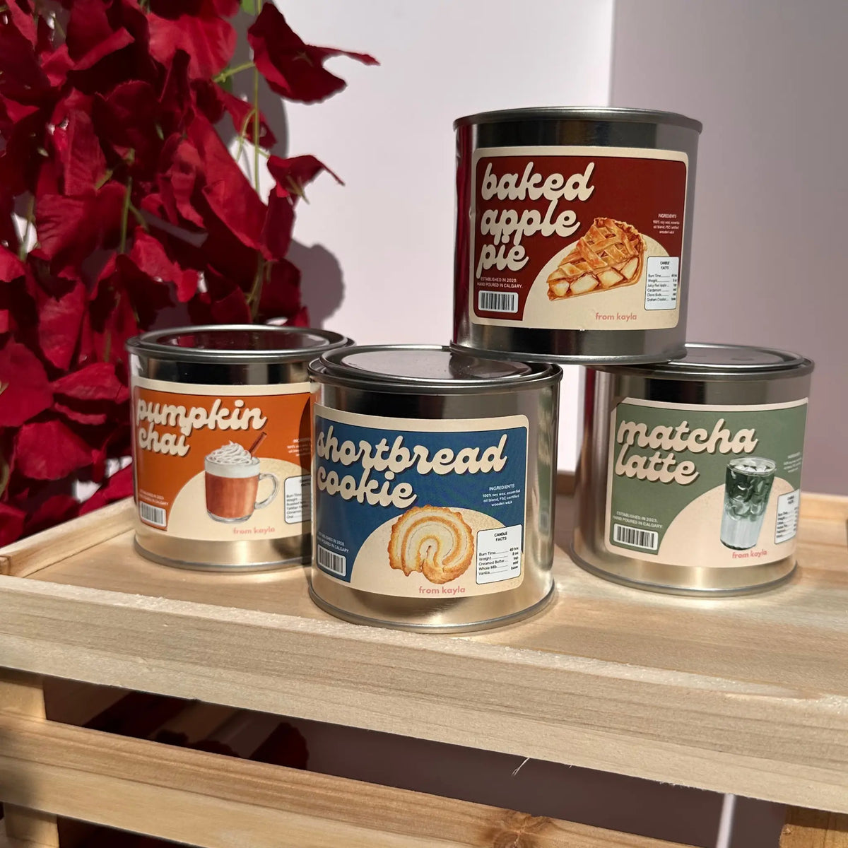 Four metal scented candles in cylindrical cans are displayed on a light wood shelf. The top can is labeled 'baked apple pie' and the three cans in front are labeled 'pumpkin chai', 'shortbread cookie', and 'matcha latte'.