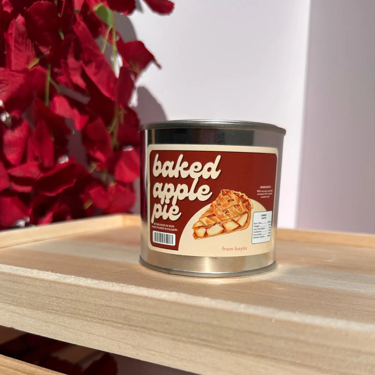A metal tin candle labeled 'baked apple pie' sits on a light wood shelf, with red decorative leaves in the background.