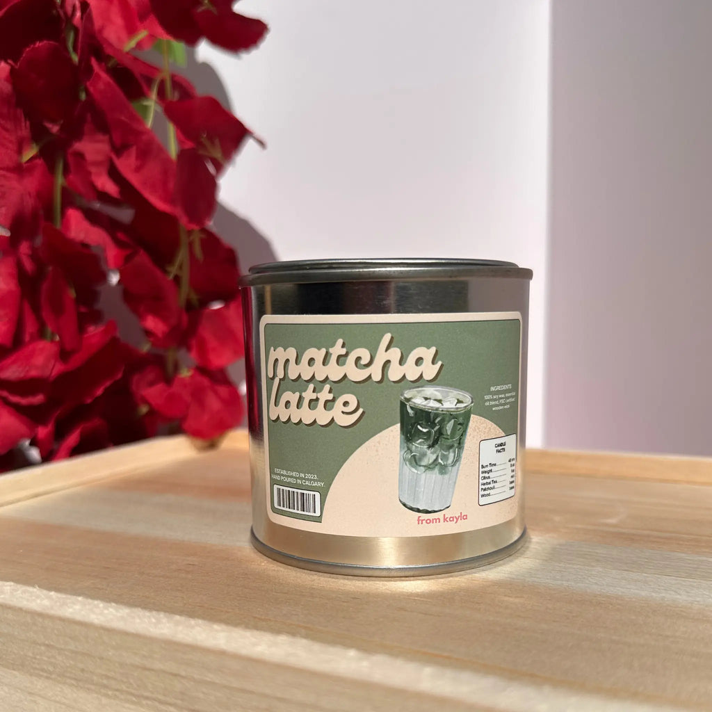 A metal tin of matcha latte coffee beans sits on a light wooden surface. The tin has a green and beige label with an illustration of a matcha latte and the words 'matcha latte' in large script.