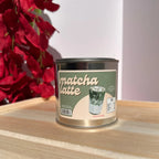 A metal tin of matcha latte coffee beans sits on a light wooden surface. The tin has a green and beige label with an illustration of a matcha latte and the words 'matcha latte' in large script.