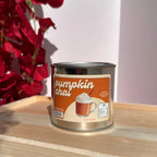 A metal tin candle labeled 'pumpkin chai' sits on a light wooden surface. The label shows an illustration of a pumpkin chai drink with whipped cream and a cinnamon stick.