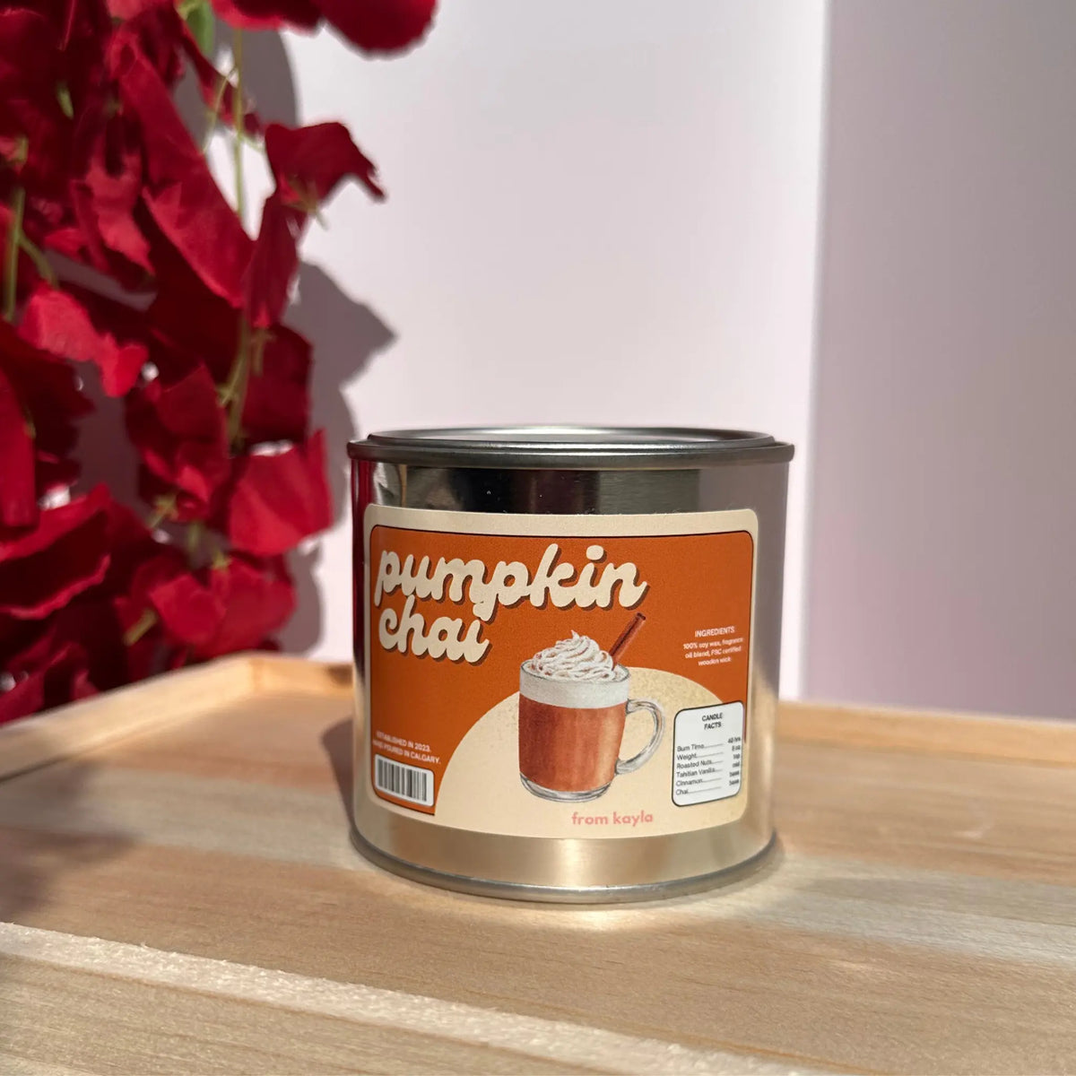 A metal tin candle labeled 'pumpkin chai' sits on a light wooden surface. The label shows an illustration of a pumpkin chai drink with whipped cream and a cinnamon stick.