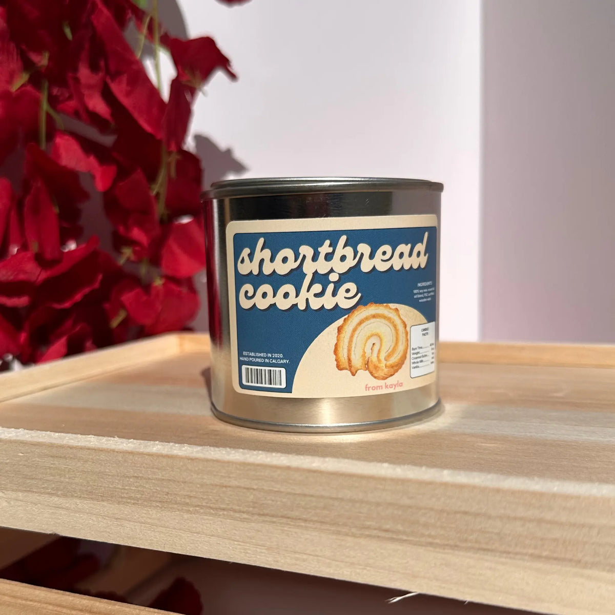 A can of 'shortbread cookie' from the brand 'from boston' sits on a light wood shelf. The can has a blue and cream label with an illustration of a shortbread roll.