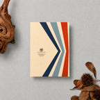 A square cream-colored greeting card or booklet with a bold chevron design in navy, light blue, and rust red, styled flat-lay on a textured gray background with dried leaves and a piece of driftwood.